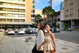 Two women are walking along a city street, with buildings and parked cars in the background. One is wearing a white blazer and black pants, while the other is dressed in a beige strapless dress. They both appear to be engaged in conversation, with a busy urban environment around them.