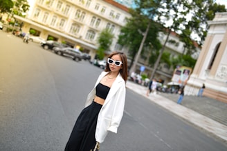 A stylish woman wearing white sunglasses, a white blazer, a black crop top, and black pants is standing prominently in an urban setting. The background features a busy street with parked cars, a historic building, and some trees.
