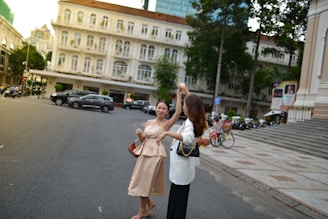Two women are engaged in a joyful dance on a city street, surrounded by classic European-style architecture. The buildings feature cream-white facades with balconies and arched windows. Trees and parked vehicles line the street, and the atmosphere is lively and carefree.