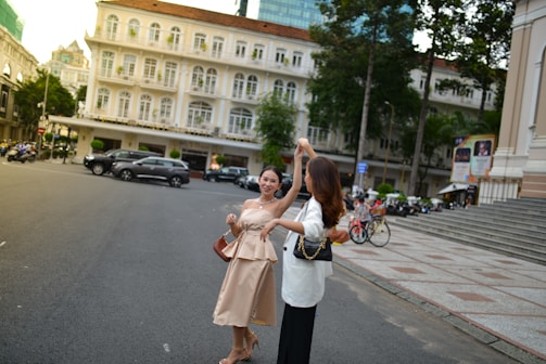 Two women are engaged in a joyful dance on a city street, surrounded by classic European-style architecture. The buildings feature cream-white facades with balconies and arched windows. Trees and parked vehicles line the street, and the atmosphere is lively and carefree.