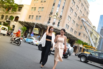 Two women walk confidently across a busy city street. One is wearing a stylish white blazer over a black outfit with sunglasses, while the other is dressed in an elegant beige dress. They are surrounded by vehicles including cars and motorcycles, and a large, upscale building prominently features a luxury brand name.