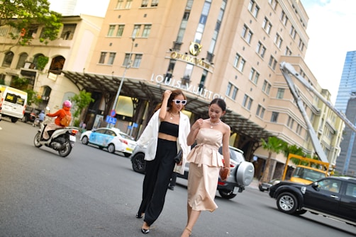 Two women walk confidently across a busy city street. One is wearing a stylish white blazer over a black outfit with sunglasses, while the other is dressed in an elegant beige dress. They are surrounded by vehicles including cars and motorcycles, and a large, upscale building prominently features a luxury brand name.