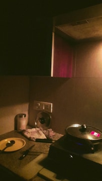 A dimly lit kitchen counter with a pot on a cooking stove. Nearby are utensils including a spoon on a yellow plate, a knife, a container, a folded cloth, a lid, and various kitchen items. An electric outlet is visible on the wall.