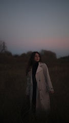 A person wearing a long coat stands in a grassy field during twilight. The sky is dim with shades of blue and pink, and the surrounding vegetation is dark and shadowy. The person's hair is down, and they appear contemplative or reflective.