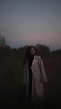 A person wearing a long coat stands in a grassy field during twilight. The sky is dim with shades of blue and pink, and the surrounding vegetation is dark and shadowy. The person's hair is down, and they appear contemplative or reflective.