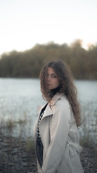 A young woman with long, curly hair stands by a lakeside in a beige trench coat. The background features a blurred view of water and trees, creating a serene and peaceful atmosphere. Her expression is introspective, with eyes looking directly at the camera. The overall setting conveys a sense of calmness and contemplation.