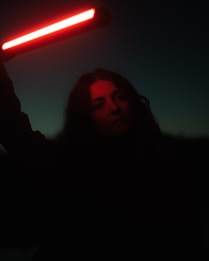 A person stands in a dark environment holding a red light stick, casting a glow on their face. The light cuts through the darkness, creating a stark contrast and highlighting the person's facial features. The background is a deep gradient from dark to less dark at the base.