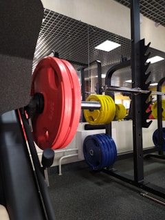 A gym setting featuring a barbell loaded with red, yellow, and blue weight plates on a squat rack. The background shows a wall mirror and ceiling lights, reflecting parts of the gym equipment.
