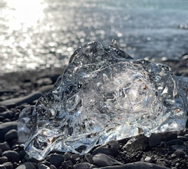A large, clear ice chunk lies on a pebble-strewn beach with sunlight glistening off the water in the background. The intricate textures and transparency of the ice are highlighted by the bright light, creating a captivating visual effect.