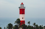 A tall lighthouse with red and white horizontal stripes stands prominently against the backdrop of the ocean. Surrounding the base of the lighthouse are green palm trees swaying gently.