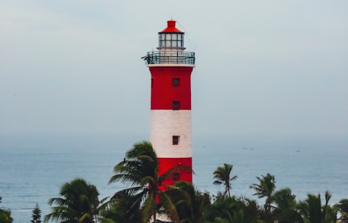 A tall lighthouse with red and white horizontal stripes stands prominently against the backdrop of the ocean. Surrounding the base of the lighthouse are green palm trees swaying gently.