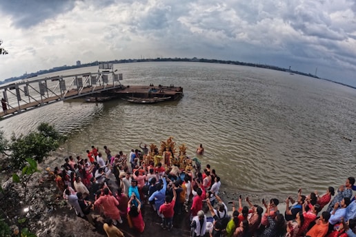 A large group of people gathered at the edge of a river, some appearing to perform a religious or cultural ritual involving colorful statues. A cloudy sky looms above, and a jetty or dock extends into the water, with a few boats moored nearby.