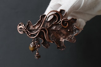 An intricate metal leaf-shaped brooch, embellished with swirling copper wire designs and small dangling beads. The brooch has a rustic, handcrafted appearance, and is being held by a gloved hand against a dark background.