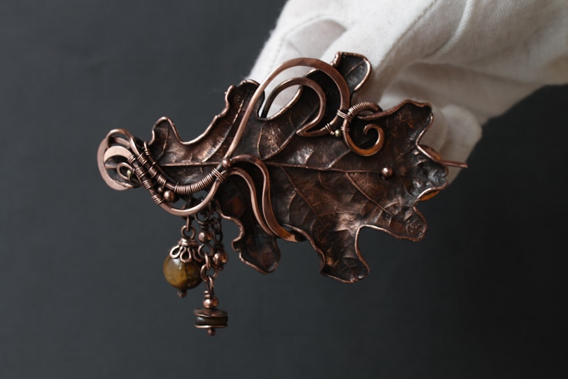 An intricate metal leaf-shaped brooch, embellished with swirling copper wire designs and small dangling beads. The brooch has a rustic, handcrafted appearance, and is being held by a gloved hand against a dark background.