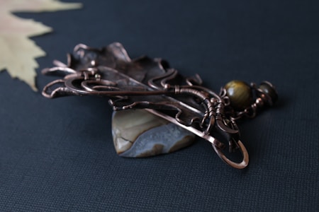 An intricately designed piece of jewelry rests on a dark fabric surface. The jewelry features a mix of metal wires and ornate elements, including a decorative stone and spherical bead. The craftsmanship highlights a blend of rustic and elegant styles.