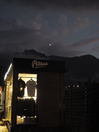 A small, illuminated shop or cabin displays hanging clothes, including jackets and shirts. The shop is situated in a dark, outdoor setting with a mountainous landscape in the background. The sky is dusky, with a visible crescent moon and scattered clouds, creating a serene ambiance.