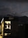 A small, illuminated shop or cabin displays hanging clothes, including jackets and shirts. The shop is situated in a dark, outdoor setting with a mountainous landscape in the background. The sky is dusky, with a visible crescent moon and scattered clouds, creating a serene ambiance.