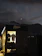 A small, illuminated shop or cabin displays hanging clothes, including jackets and shirts. The shop is situated in a dark, outdoor setting with a mountainous landscape in the background. The sky is dusky, with a visible crescent moon and scattered clouds, creating a serene ambiance.