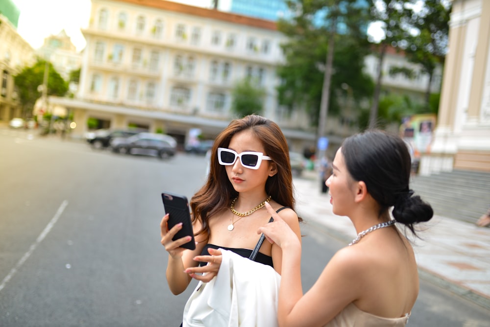 two women standing on the side of a road looking at a cell phone
