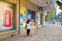 Two people, one wearing a white shirt and holding a bag and the other in a beige dress with a hat, walk arm-in-arm down a sidewalk beside a building with colorful travel-themed posters. A sign for a Tourist Information Center hangs above.