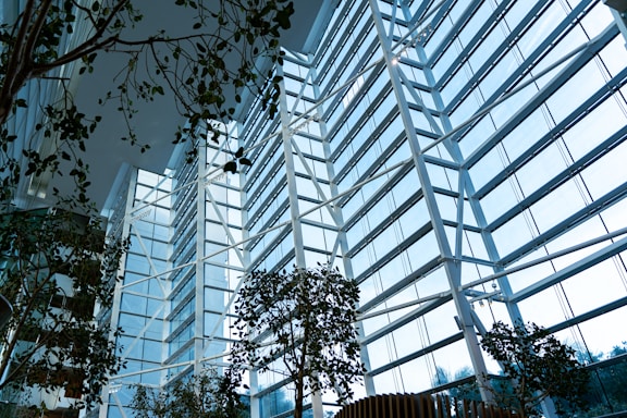 A modern, spacious interior filled with natural light streaming through large, floor-to-ceiling glass windows. The structure is characterized by tall white beams and steel framework. Several lush green plants are arranged throughout the space, contributing to a harmonious blend of nature and architecture.