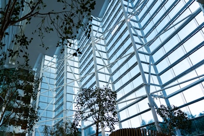 A modern, spacious interior filled with natural light streaming through large, floor-to-ceiling glass windows. The structure is characterized by tall white beams and steel framework. Several lush green plants are arranged throughout the space, contributing to a harmonious blend of nature and architecture.