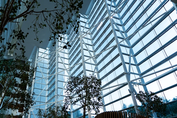 A modern, spacious interior filled with natural light streaming through large, floor-to-ceiling glass windows. The structure is characterized by tall white beams and steel framework. Several lush green plants are arranged throughout the space, contributing to a harmonious blend of nature and architecture.