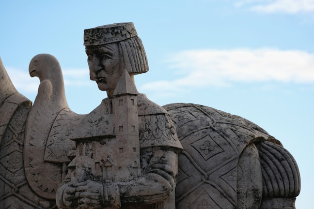 A stone sculpture depicting a historical figure adorned with a headdress and intricate clothing, holding a model of a castle. A bird with an extended neck is carved into the background, and a large, patterned shield is partially visible behind the figure.
