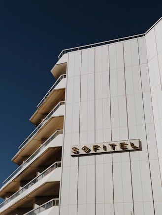 The image depicts a multi-story building with a modern architectural design. The structure features multiple balconies on one side and is marked with the name 'SOFITEL' in distinct letters on the front facade. The sky in the background is clear and deep blue, contrasting with the light beige color of the building.