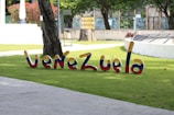 Colorful 3D letters spelling 'Venezuela' are placed on a grassy area in an outdoor setting. Trees and a concrete walkway are visible in the background along with some signage and a white ornamental structure.