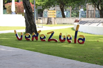 Colorful 3D letters spelling 'Venezuela' are placed on a grassy area in an outdoor setting. Trees and a concrete walkway are visible in the background along with some signage and a white ornamental structure.