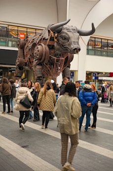 A large mechanical bull sculpture is surrounded by a crowd of people inside a modern indoor venue. The bull is crafted from metal with visible gears and intricate details. People in winter clothing are gathered around, some observing the sculpture closely.