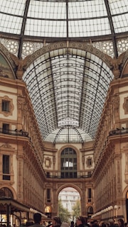 A large, ornate shopping arcade with a glass dome ceiling adorned with intricate metalwork and a combination of classical and neoclassical architectural details. The interior is bustling with people walking beneath the high, arched ceilings and along the elegant corridors.