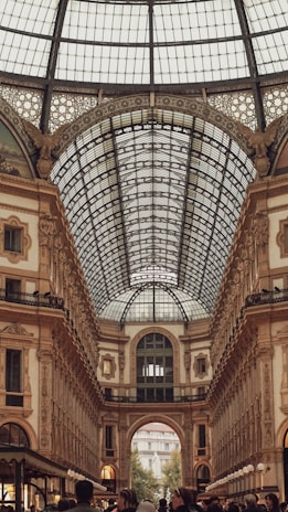 A large, ornate shopping arcade with a glass dome ceiling adorned with intricate metalwork and a combination of classical and neoclassical architectural details. The interior is bustling with people walking beneath the high, arched ceilings and along the elegant corridors.