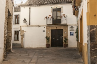 A quaint, cobblestoned street corner with a traditional whitewashed building featuring a wrought iron balcony and a dark wooden door. Two potted plants flank the entrance. The walls have decorative tiles and a vintage lamp. The architecture reflects a rustic, historical charm.