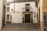 A quaint, cobblestoned street corner with a traditional whitewashed building featuring a wrought iron balcony and a dark wooden door. Two potted plants flank the entrance. The walls have decorative tiles and a vintage lamp. The architecture reflects a rustic, historical charm.