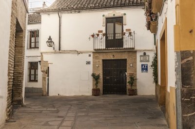 A quaint, cobblestoned street corner with a traditional whitewashed building featuring a wrought iron balcony and a dark wooden door. Two potted plants flank the entrance. The walls have decorative tiles and a vintage lamp. The architecture reflects a rustic, historical charm.