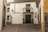 A quaint, cobblestoned street corner with a traditional whitewashed building featuring a wrought iron balcony and a dark wooden door. Two potted plants flank the entrance. The walls have decorative tiles and a vintage lamp. The architecture reflects a rustic, historical charm.