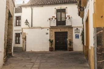 A quaint, cobblestoned street corner with a traditional whitewashed building featuring a wrought iron balcony and a dark wooden door. Two potted plants flank the entrance. The walls have decorative tiles and a vintage lamp. The architecture reflects a rustic, historical charm.