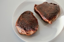 Two pieces of seared meat resting on a white plate. The steaks have a dark, caramelized crust and appear cooked with a juicy interior.