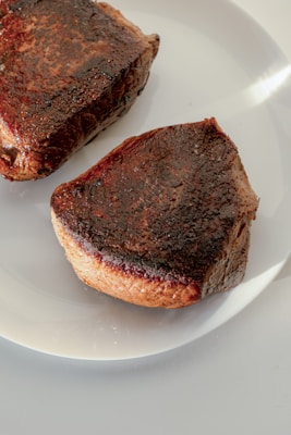 Two pieces of seared steak are resting on a white plate. The meat has a rich, dark brown crust with visible grill marks, indicative of being cooked to a perfect sear. The surface glistens slightly, suggesting juiciness and flavor.