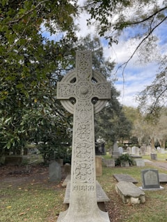 A detailed stone cross stands in the foreground, intricately carved with circular and floral patterns. It has the name 'GUERRY' inscribed at the base. The background features a graveyard with several tombstones surrounded by trees, some with sparse foliage. The sky appears partly cloudy, casting a serene atmosphere over the scene.