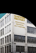 A multi-story industrial building with large grid-patterned windows and a painted sign on the facade. The text on the building reads 'Custard Factory' followed by the phrases 'Industrious, Individual, In It Together' in bold yellow and black letters. The building is white and situated against a clear blue sky.