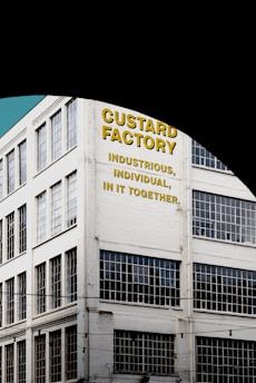 A multi-story industrial building with large grid-patterned windows and a painted sign on the facade. The text on the building reads 'Custard Factory' followed by the phrases 'Industrious, Individual, In It Together' in bold yellow and black letters. The building is white and situated against a clear blue sky.