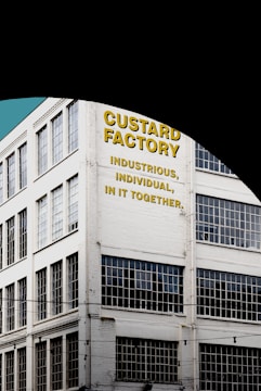 A multi-story industrial building with large grid-patterned windows and a painted sign on the facade. The text on the building reads 'Custard Factory' followed by the phrases 'Industrious, Individual, In It Together' in bold yellow and black letters. The building is white and situated against a clear blue sky.