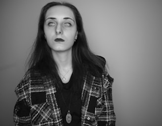 A person with long dark hair, pale skin, and dark lipstick is wearing a plaid jacket over a dark top. The person also has visible piercings and appears to be wearing a pendant necklace. The background is plain and the image is in grayscale with a focus on the eyes.