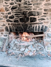 A rustic stone fireplace with a small fire burning at the base. The stones are dark with mortar, and the fire emits bright orange and yellow flames. Pieces of wood and ash are scattered around the base.