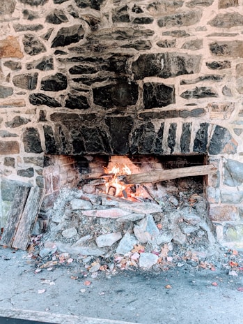 A rustic stone fireplace with a small fire burning at the base. The stones are dark with mortar, and the fire emits bright orange and yellow flames. Pieces of wood and ash are scattered around the base.