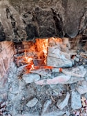 A rustic stone fireplace with a vibrant fire burning, surrounded by ash and charred logs. The interior is lined with bricks and the stone exterior appears weathered and rugged.