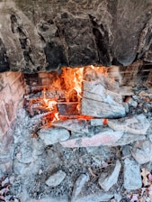 A rustic stone fireplace with a vibrant fire burning, surrounded by ash and charred logs. The interior is lined with bricks and the stone exterior appears weathered and rugged.